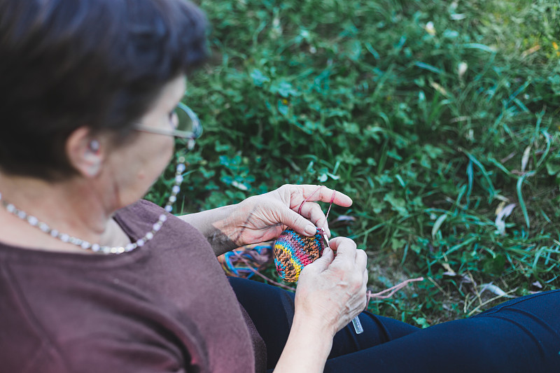 Rear,view,of,senior,woman,knitting,in,nature,on,a,summer,day,–,Pretty,old,lady,with,brown,hair,sitting,on,the,grass,while,making,crochet,clothes