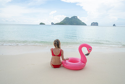 年轻女子放松在田园诗般的海滩上坐在充气火烈鸟在泰国的岛屿。人们旅游目的地有趣和酷的态度概念