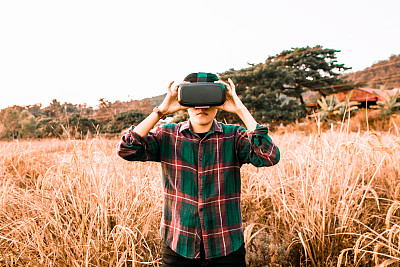 一名亚洲男子戴着一副虚拟现实眼镜在金色稻田里融化成像素。未来科技与自然概念