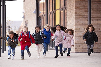 一个快乐的多民族的年轻学校的孩子们穿着大衣，背着书包和他们的同学在他们的幼儿学校大楼外面的人行道上跑步