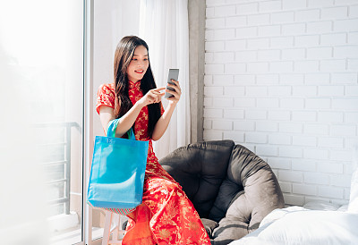 肖像的微笑美丽的亚洲女人穿着中国服装，旗袍，旗袍春节使用智能手机技术购物和拿包坐在干净的白色房间，复印空间靠窗。