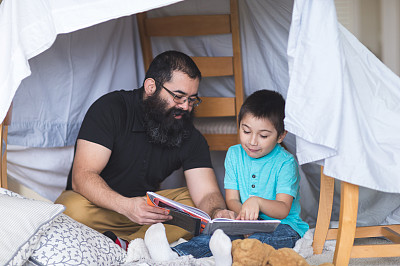 美国土著爸爸和他的小儿子在客厅里临时搭建的堡垒下读书