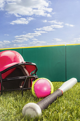 一个黄色和粉色的垒球，一个铝制的球棒和粉色的击球手的头盔，放在有阳光的人造草坪上