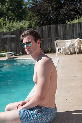 一个健康的男人坐在泳池边，脚在水里，没穿衬衫，穿蓝色泳裤，戴着飞行员太阳镜。一个白人站在后院的泳池边，看起来很严肃。