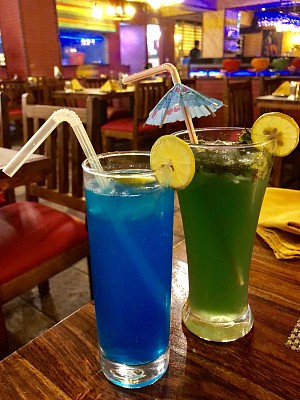 “蓝礁湖”酒精鸡尾酒和维珍莫吉托非酒精鸡尾酒在高球杯与橙色装饰和吸管，印度酒吧背景照片
