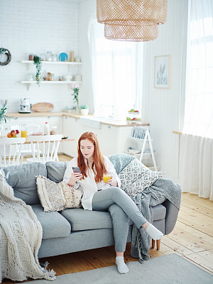 漂亮的红头发年轻女子坐在家里的沙发上看手机短信，看起来很惊讶。她手里拿着一杯橙汁