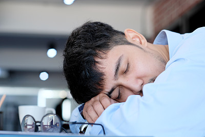 疲惫的亚洲人在办公桌前睡觉。戴着眼镜的年轻商人过度劳累而睡着了，创造性的休闲男人在他的工作场所睡觉