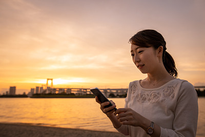 一名年轻女子在东京湾日落海滩上使用智能手机