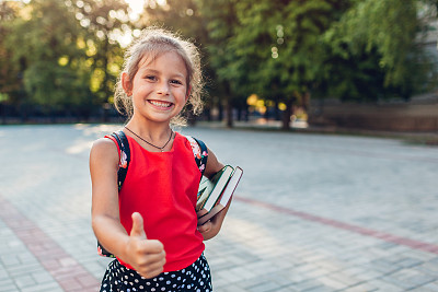 快乐的小学生女孩背着书包，拿着书。小学生在户外竖起大拇指。教育