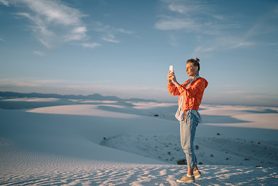 年轻女子在白沙沙漠用智能手机拍摄以风景为背景的自拍，时髦女孩站在沙丘上欣赏美国国家纪念碑的美丽风景