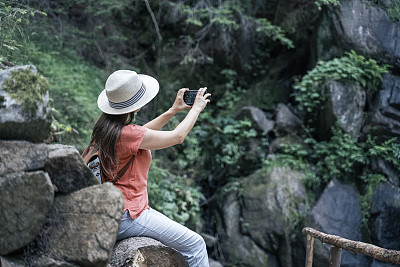 回归自然。独自旅行者在瀑布附近用智能手机拍照。一个快乐的年轻女游客漫步在大自然中。阳光明媚的日子里在森林里的户外享受。