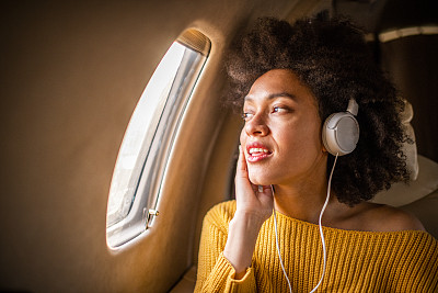 年轻时尚的女人坐在私人飞机上，看着窗外，听着耳机里的音乐