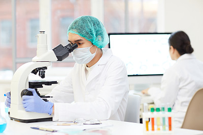 严肃专注的年轻亚洲男性科学家戴着无菌帽和口罩坐在桌子旁，通过显微镜分析实验样品
