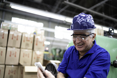 快乐的工程师拿着智能手机和使用防护服在工厂工作
