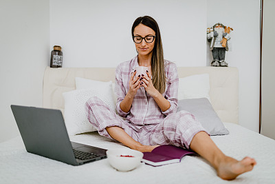 穿着睡衣在床上喝咖啡用笔记本电脑的女人