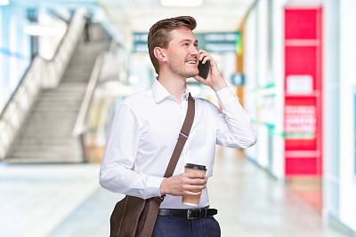 年轻男性专业人士站在办公室散焦背景前，戴着邮差包，拿着包，使用手机