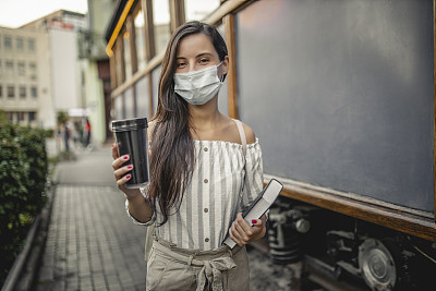 带着医用口罩的妇女在户外散步，喝咖啡，拿着书