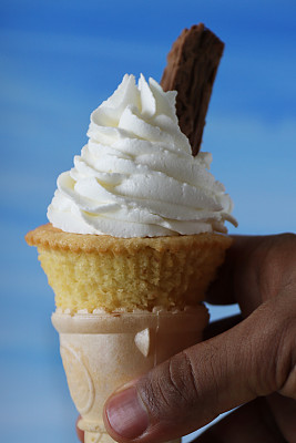 特写:一个不知名的人拿着冰淇淋甜筒设计的纸杯蛋糕，自制香草蛋糕上面覆盖着管状糖霜和巧克力片，蓝天背景，关注前景