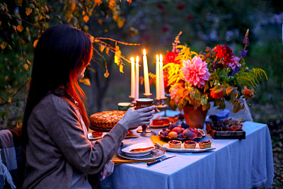 秋夜摄影:年轻女子与酒杯在黄昏的自然户外浪漫晚餐。餐桌装饰-蛋糕，无花果，盘子，烛台蜡烛，落花。红色的大丽花花束