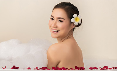 放松美丽的亚洲女人纵容她的身体在温泉浴缸与白色泡沫泡沫的肥皂和玫瑰花瓣。休闲的追求