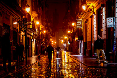 西班牙巴伦西亚，雨后，当地人在被橙色灯光照亮的“Carrer de la Bosseria”街道上散步或骑车