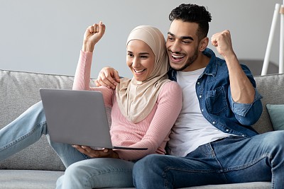 一对情绪激动的阿拉伯夫妇拿着笔记本电脑举起拳头庆祝成功
