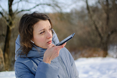 一位穿着蓝色外套的女士正在发送语音信息，解释在室外与移动助手通话的智能手机上的一些事情。用智能手机语音识别和听写的女孩