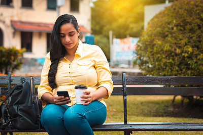 年轻的拉丁女人在公园喝咖啡时查看手机