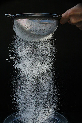 一个不认识的人的手拿着一个金属筛子，白色的面粉被筛在黑色背景下，白色的粉末落在半空中，聚焦前景