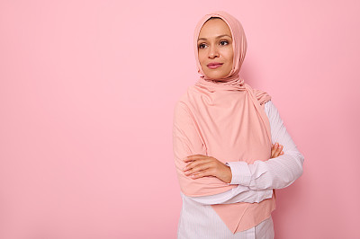 自信的阿拉伯穆斯林美丽女人的肖像，有迷人的目光和眼神，用粉红色的头巾盖住头部，站在彩色背景的四分之三复制空间，望向别处