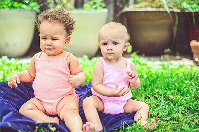 两个婴儿一起坐在外面，他们穿着相同的衣服，年龄相同，但大小和种族不同