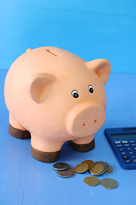 近景的小猪储蓄罐坐在计算器和成堆的硬币，蓝色背景，聚焦前景，抵押，家庭财务和储蓄的概念