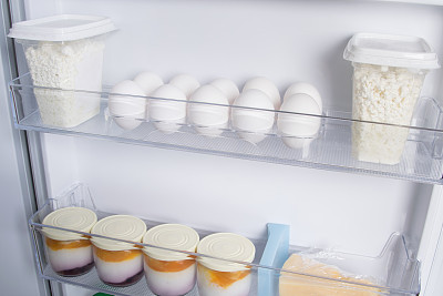 在白色冰箱的门上，架子上放着鸡蛋、奶酪、酸奶和白软干酪