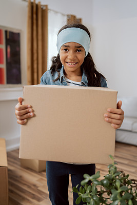 一个甜美、漂亮的学龄女孩带着温柔的微笑看着镜头，为搬家感到高兴，带着打包好的轻纸盒走出家门，帮助她的父母