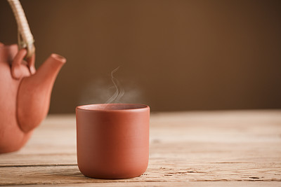 棕色陶瓷杯配茶或咖啡和热蒸汽，在质朴的木桌上，茶壶或咖啡壶的局部视野，棕色背景，文字空间。