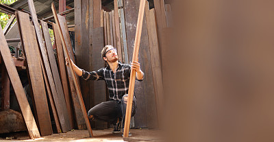年轻的白人木匠正在选择木板在他自己的车库风格的工作室与复制空间的爱好