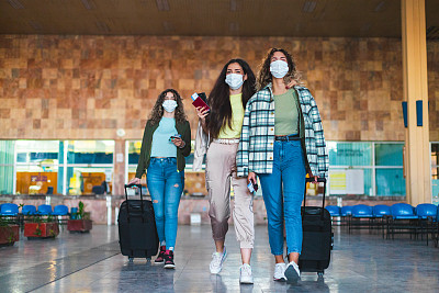 三位女性朋友戴着防护口罩，拿着护照和机票，拖着行李箱在机场行走