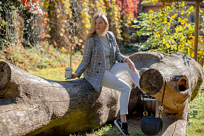 一位中年妇女，一头金黄色长发，穿着格子夹克和卷领毛衣，坐在干燥的木材上，手里拿着咖啡。