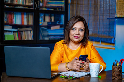 Happy middle-aged businesswoman or teacher in glasses sit at home office typing on laptop and using mobile phone.