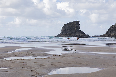 两个带着冲浪板的人走在安静的海滩上，远处有岩石，天空多云