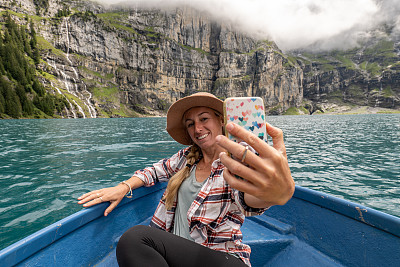 年轻女子在划艇上自拍，而巡航在瑞士阿尔卑斯山的高山湖泊。人们户外旅行的乐趣概念