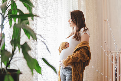 幸福的孕妇站在窗边，抱着她的肚子