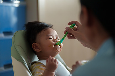 坐在高椅子上的亚洲小男孩正在被妈妈喂食。