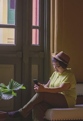 快乐的亚洲成年女性游客在垂直框架的老式建筑内的沙发上放松时使用智能手机