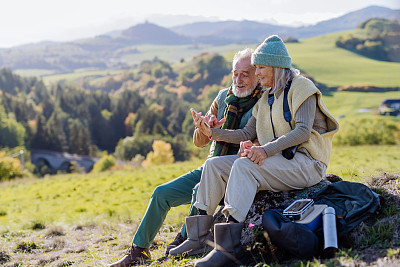 在秋天的大自然中，一对老年夫妇在远足时休息，看手机。
