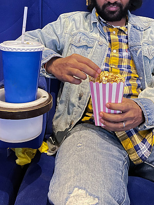 特写图像的印度男子坐在一排电影院座位，拿着硬纸盒焦糖爆米花，一次性饮料杯与吸管在饮料架，重点在前景