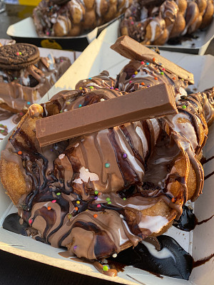 硬纸板箱里的泡泡华夫蛋筒的特写图像，冰淇淋上覆盖着巧克力布朗尼块，糖屑，焦糖太妃酱，饼干，糖果，巧克力糖浆，在街头食品市场的摊位店供应，重点在前景