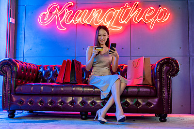 肖像照片的年轻美丽的亚洲女模特穿着优雅的衣服在霓虹灯的房间支付与她的信用卡借记卡在她的手机网上购物狂欢
