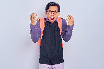 愤怒的年轻亚洲学生男子穿着休闲服装，背着双肩包，耸肩，在灰色背景上分开双手。高中大学学院概念