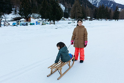 大一点的孩子和小一点的孩子一起拉雪橇，背景是美丽的冬天的山